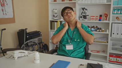 Young man in medical scrubs sits indoors in a clinic office, appearing stressed, with a stethoscope around his neck and a wheelchair in the background.