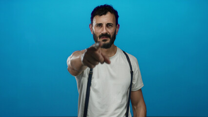 Fototapeta premium Hispanic man with beard pointing forward in blue background wearing white shirt and suspenders, conveying focus and direction in a modern setting.