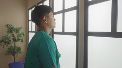 Young chinese doctor in green scrubs stands thoughtfully in a hospital clinic room interior gazing...