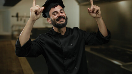 Hispanic male chef with beard pointing upwards in a professional kitchen, showcasing enthusiasm and...