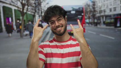 Young man wearing glasses and striped shirt flashing rock gesture on busy urban street with blurred...