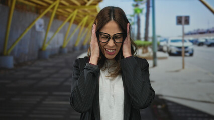 Fototapeta premium Hispanic woman on a city street feeling stressed with eyes closed and mouth clenched, surrounded by urban environment and cars.