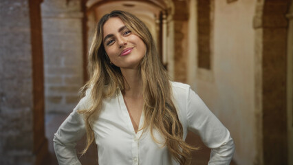 Fototapeta premium Blonde woman standing with arms on hips wearing white shirt under an arched walkway at a university campus; confidence ambition enthusiasm empowerment.