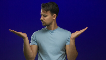 Young hispanic man in a light blue shirt extends hands to both sides while pondering a decision...