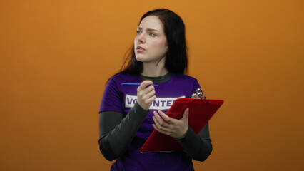 Woman volunteer with brunette hair writing on clipboard with pen against vibrant orange background...