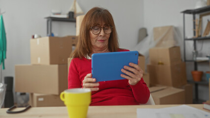 Woman in red sweater having a video call with a tablet in a new home surrounded by moving boxes and a yellow mug on the table in a cozy living room.