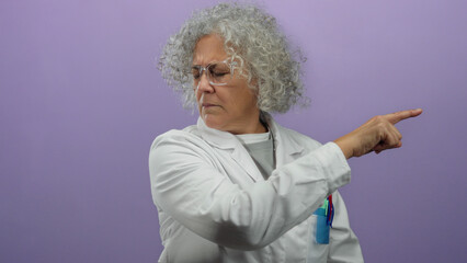 Woman pointing against a purple background wears a white lab coat displaying a serious expression...