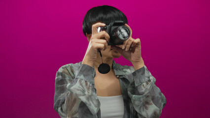Woman holds camera against pink background wearing camo jacket showcasing photography hobby with confident expression.