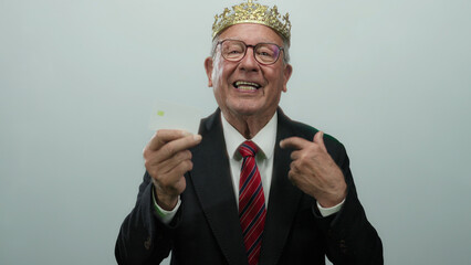 Senior man wearing a suit and crown presents a credit card with a confident expression against a...