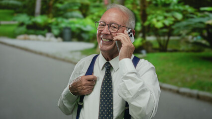Senior man in suit talking on smartphone, smiling outdoors in lush green park environment,...