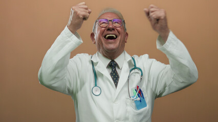 Senior male doctor in white uniform celebrates with raised hands against an isolated brown...