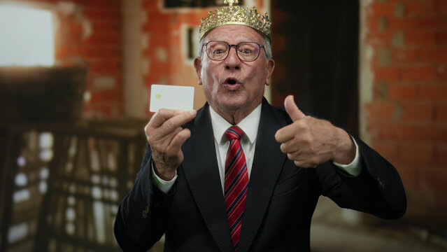 Elderly man in suit and crown gives thumbs up, holding card at indoor construction site, embodying business and triumph.