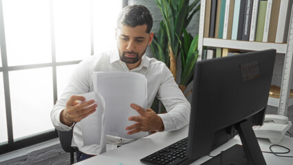 Young man reading documents at desk in modern office with computer and bookshelf nearby, showcasing...