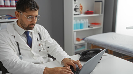 Young man wearing lab coat and glasses works on laptop in clinic room with medical equipment...