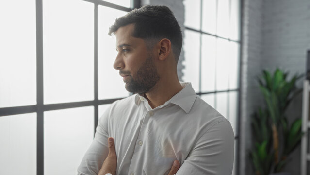 Young man looking through office window with arms crossed, appearing thoughtful and reflective in a modern indoor workplace setting, sunlight illuminating his profile.
