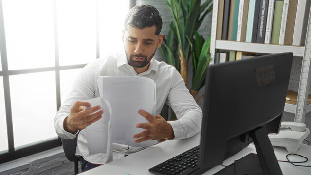 Young man reading documents at desk in modern office with computer and bookshelf nearby, showcasing a professional work environment under soft natural light. - Powered by Adobe