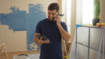 Young man smiling while talking on phone and holding tablet in living room of new home during renovation with paint supplies scattered around.