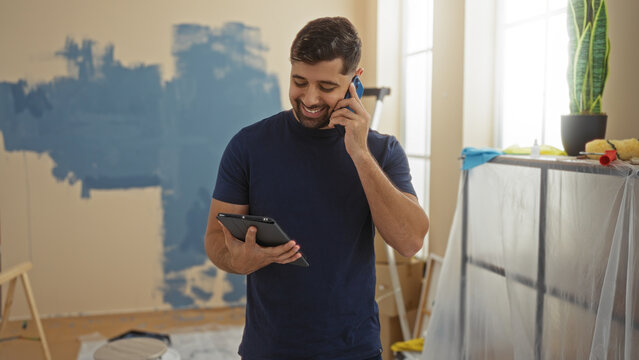 Young man smiling while talking on phone and holding tablet in living room of new home during renovation with paint supplies scattered around.