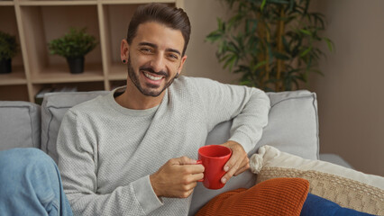 Young hispanic man smiling with a red mug in a cozy living room, surrounded by pillows and plants, showcasing a relaxed indoor ambiance.