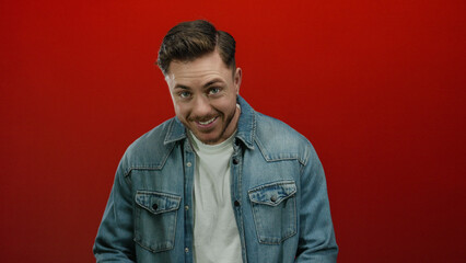 Young man with beard in denim shirt stands against vibrant red background looking contemplative