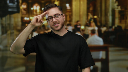 Young man with beard making gesture in a church interior, wearing glasses and black shirt, smiles...