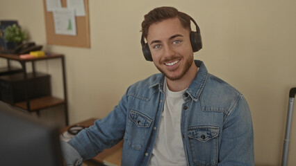 Young man with beard wearing headphones in an office setting, focused on work at computer in an indoor workplace environment, featuring casual attire and modern equipment.