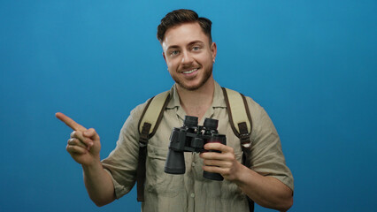 Young man with beard and backpack holding binoculars smiles and points against a blue background,...