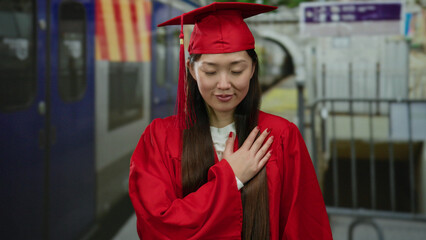 Woman in red graduation gown stands confidently near a train station backdrop, conveying pride and...