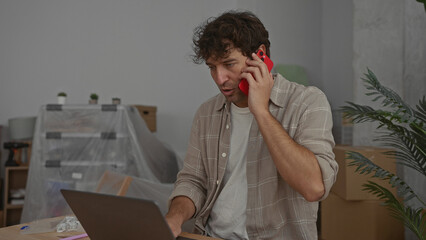 Man talking on phone while using laptop in new home filled with unpacked boxes and wrapped...