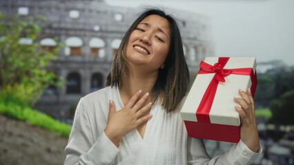 Young woman holding gift box with red ribbon in front of roman coliseum showing excitement and...