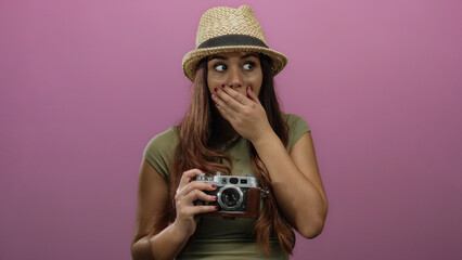 Young woman wearing a hat holding a vintage camera over a pink background with a surprised...