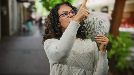 Woman holding american dollars on city street points at direction with serious expression outdoors