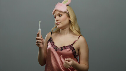 Obraz premium Young blonde woman in sleepwear holding toothbrush against grey background wearing pink sleep mask and examining toothbrush closely