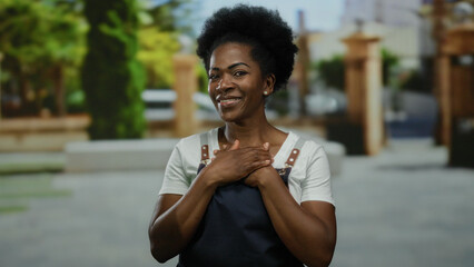 Woman smiling outdoors with hands on chest, wearing an apron, in a city street setting, embodying a...