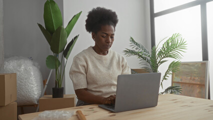 Woman working on laptop surrounded by moving boxes and plants in a new home living room suggesting...