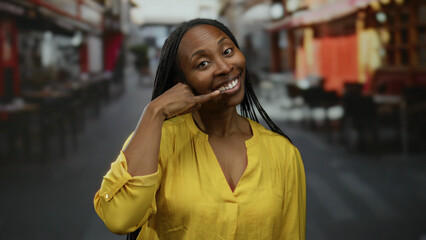 Woman wearing yellow shirt gesturing phone call while smiling in an urban street setting,...