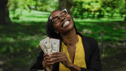 Woman outdoors holding american dollars happily with a vibrant background of a green park showing...