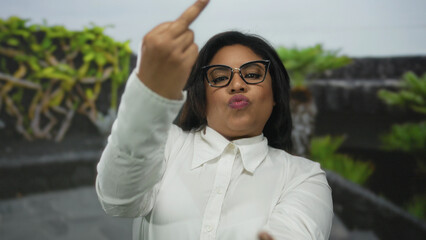 Young latin woman in white shirt makes rude gesture with middle fingers in a park setting showing...