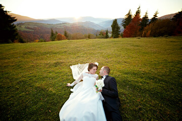 A bride and groom are lying on the grass, with the bride holding a bouquet. The scene is serene and...