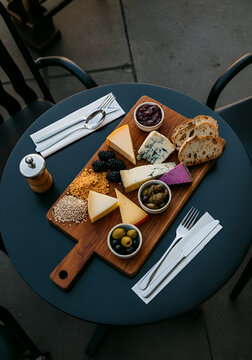  gourmet cheese platter with assorted colorful cheeses, seeds, jam, blackberries, olives, pickled vegetables, rustic bread and elegant cutlery, arranged on a dark modern table at an outdoor caf&eacute;. 