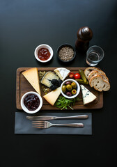  gourmet cheese platter with assorted colorful cheeses, seeds, jam, blackberries, olives, pickled vegetables, rustic bread and elegant cutlery, arranged on a dark modern table at an outdoor caf&eacute;. 