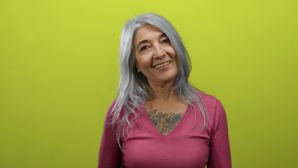 Senior woman with gray hair and tattoo wearing pink sweater smiles warmly against a vibrant yellow...