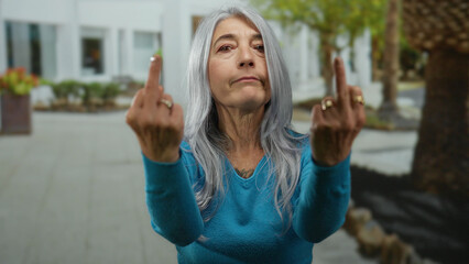 Senior woman with grey hair gesturing with middle fingers in urban outdoor setting wearing blue top.