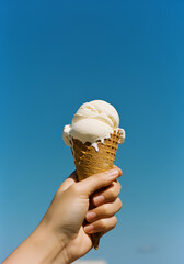 A close-up photo of a hand holding a melting vanilla ice cream cone against a clear blue sky. The ice cream is dripping down the cone and fingers, with creamy textures and warm sunlight.