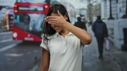 Woman covering face with hand standing on busy city street with iconic red bus in background,...