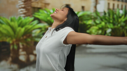 Young latina woman joyfully stretches her arms in a vibrant outdoor setting, exuding happiness and...