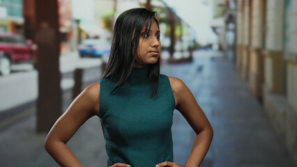 Woman standing confidently on an urban street, showcasing a thoughtful expression with blurred background and casual attire, embodying a young, independent, hispanic identity outdoors.