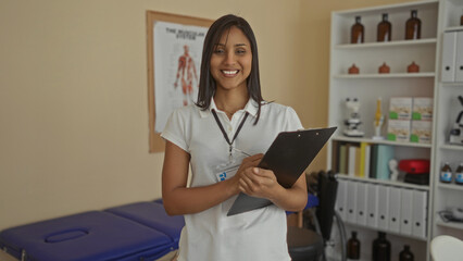 Woman smiling in a clinic taking notes on a clipboard wearing a badge standing near a poster with medical equipment in the background.
