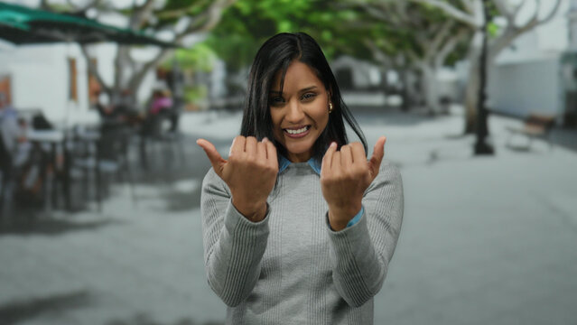 Woman smiling making come here gesture on a tree-lined street, engagingly inviting outdoors during daytime, wearing casual grey sweater in urban environment.