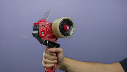 Man holds red tape dispenser against purple wall showing gripping hand with a firm grasp, highlighting packaging with an isolated background in a vibrant and focused scene.